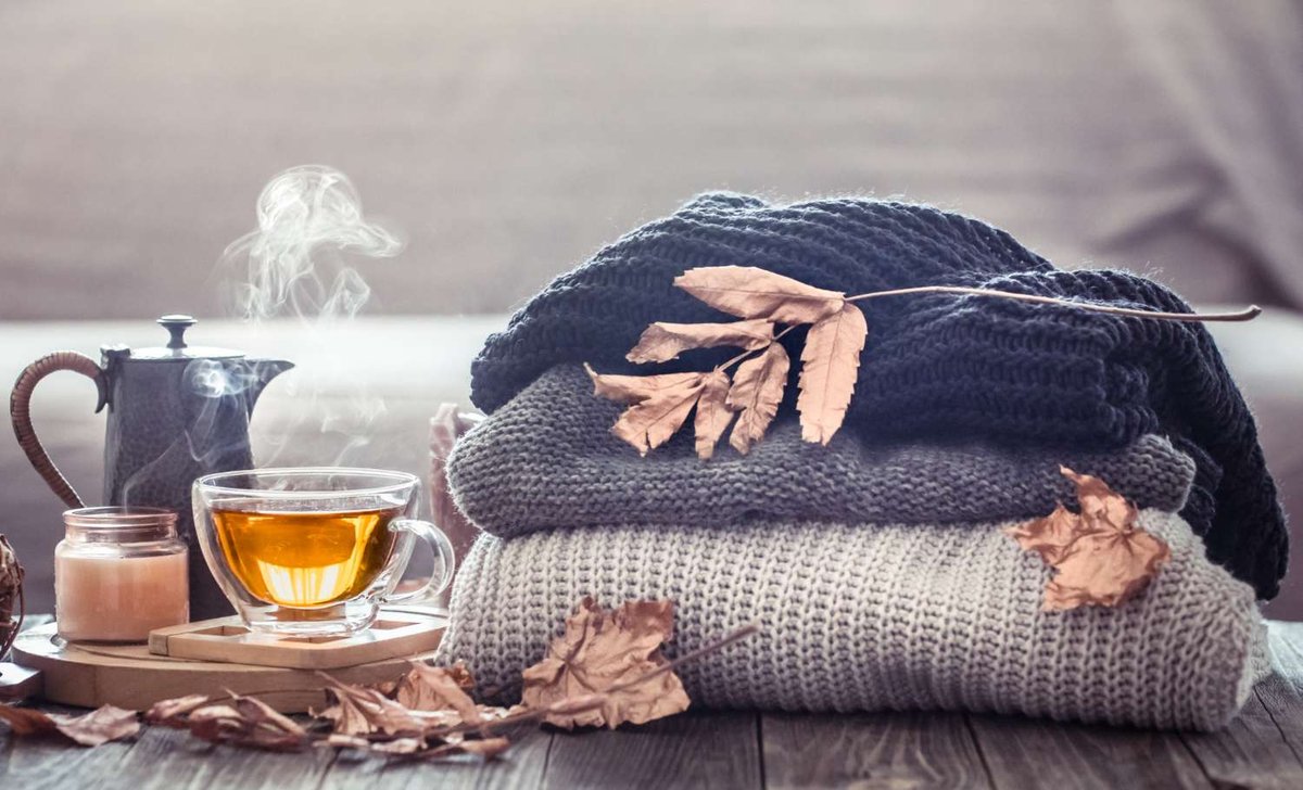 A short caption: folded wool sweaters, dried autumn leaves, and a steaming cup of tea beside a candle set a cozy October travel-packing mood.