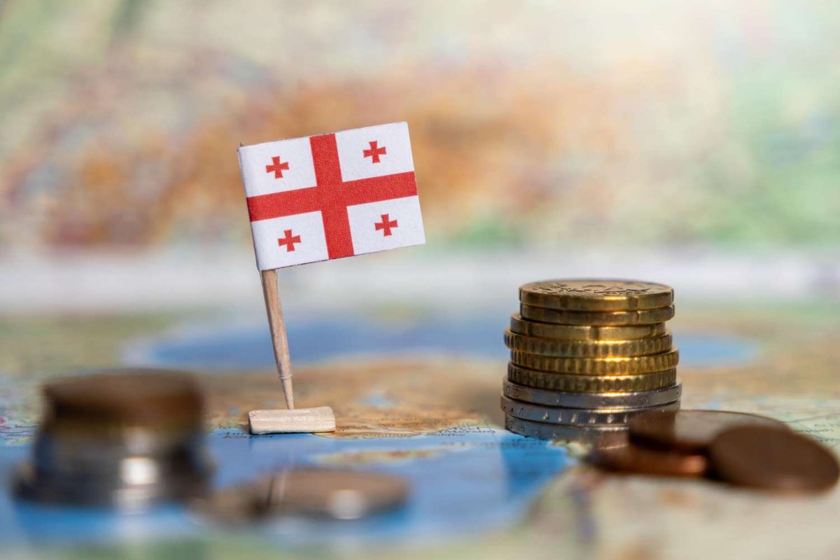 A small Georgian flag on a pin stand beside stacks of coins arranged on a colorful world map.