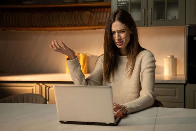 A young woman with a furrowed brow looks at her laptop in confusion as she reads a negative review.