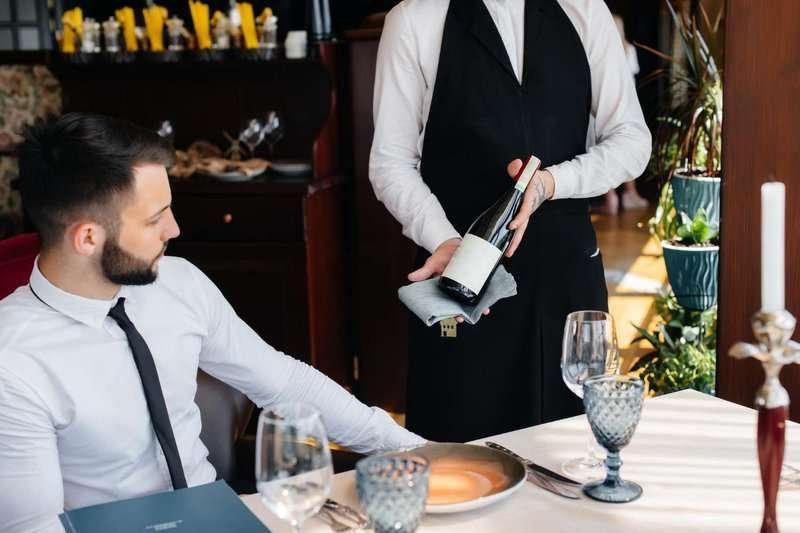 A young waiter in a stylish apron demonstrates and offers a customer an exquisite wine in a restaurant