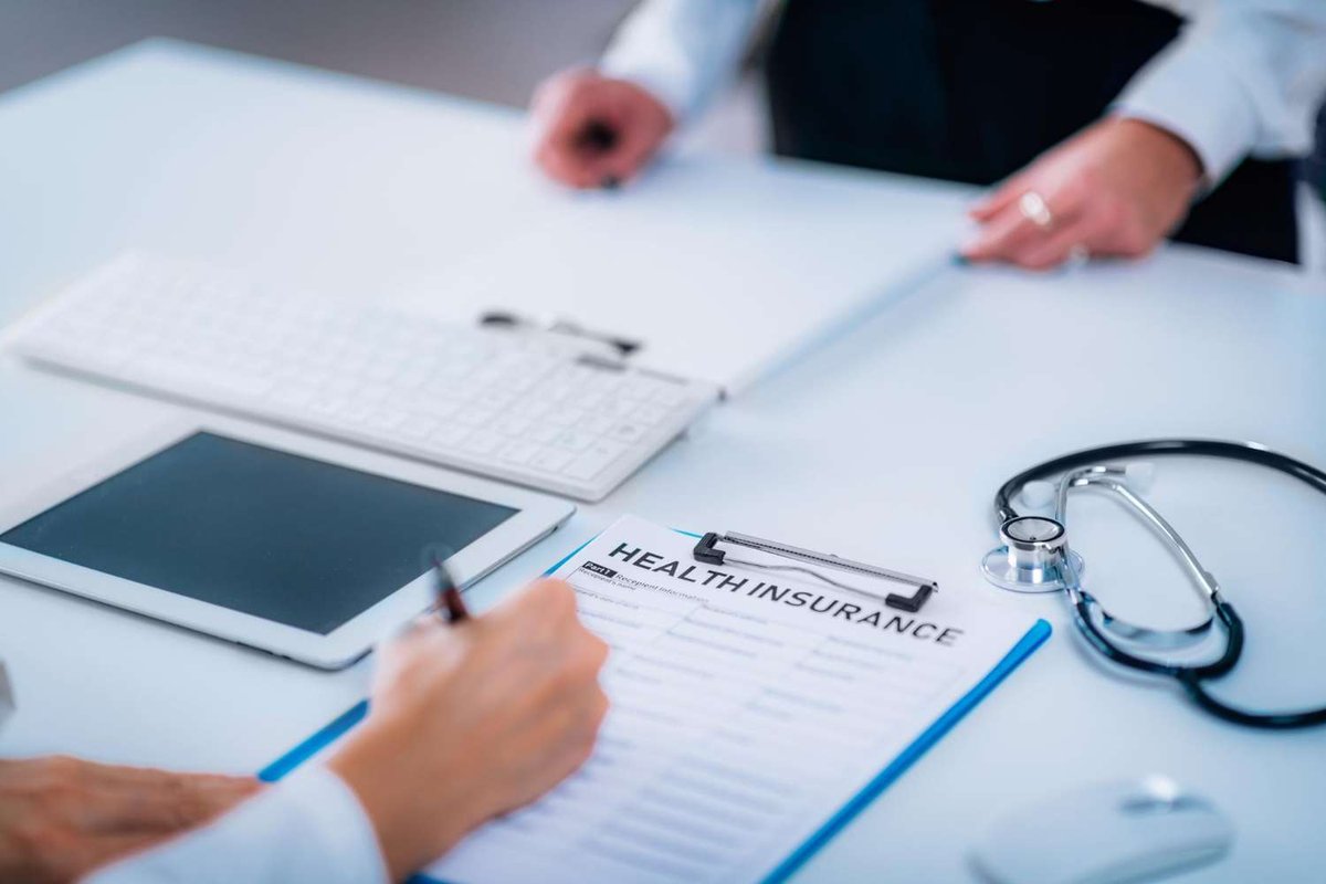 A hand fills out a health insurance form beside a stethoscope, illustrating the paperwork behind choosing between resident and tourist medical coverage.