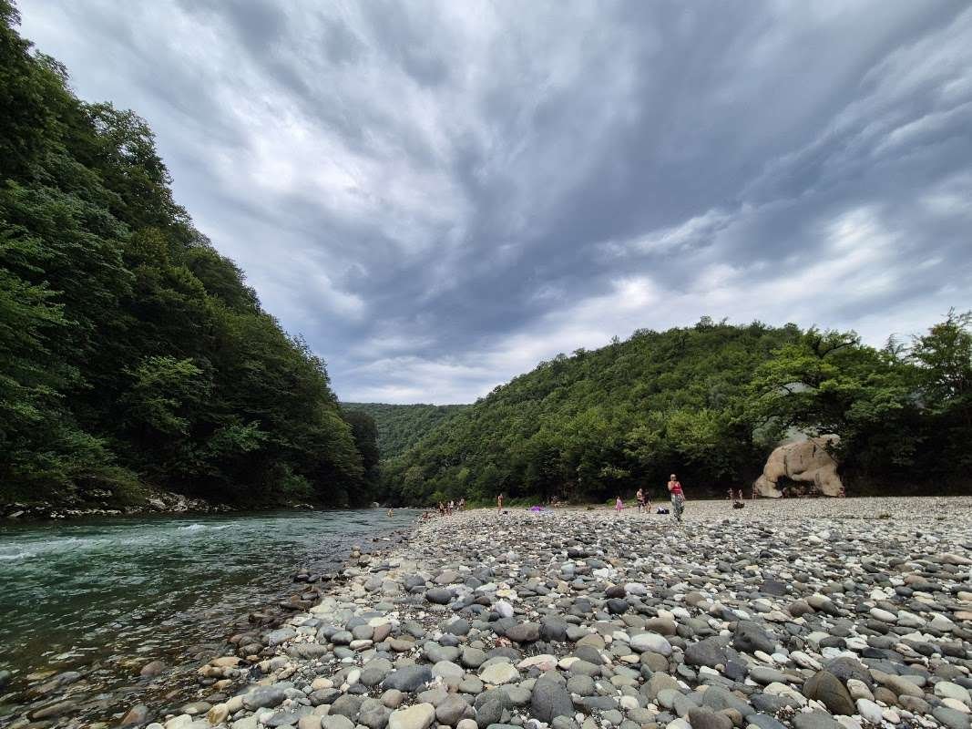 People wading and relaxing on a wide pebble beach along the Techuri river, with forested hills and a cloudy sky above.