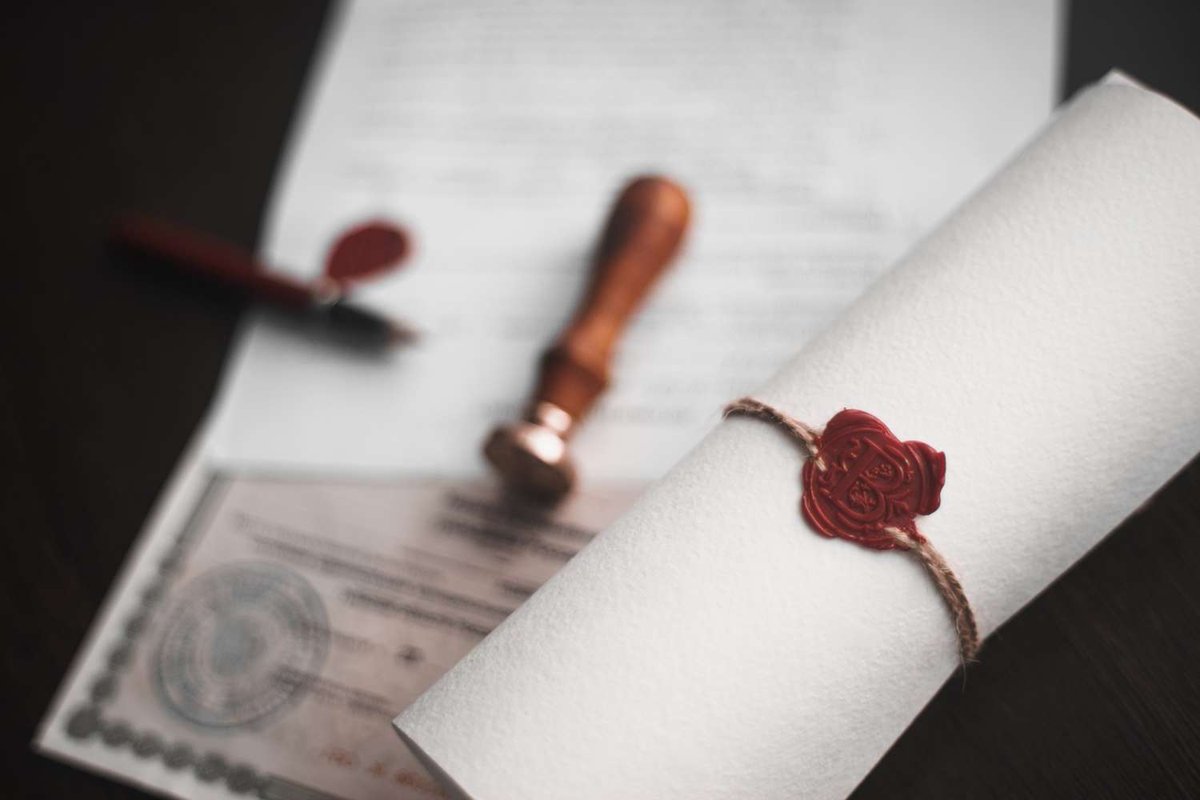 A rolled paper tied with string and sealed in red wax, next to a certificate and a wooden notary stamp on a dark desk.