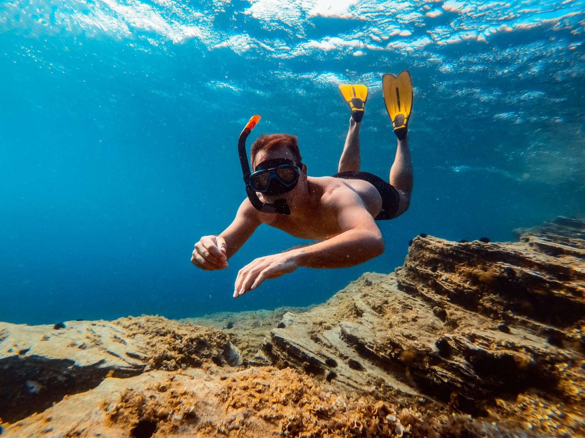 A man in a snorkel mask and yellow fins swimming over a rocky underwater reef in clear blue water.
