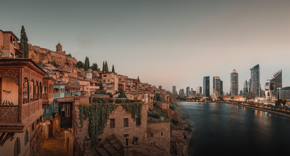 Carved wooden balconies of Old Tbilisi lean over the Kura river at dusk, with the city's glass towers and ferris wheel lit up on the opposite bank.