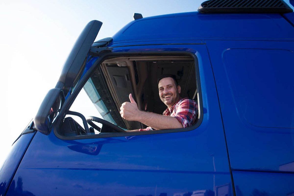 A smiling truck driver giving a thumbs up out of his blue truck's window.